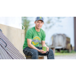 Un hombre sentado con una camiseta verde de John Deere, una gorra gris y verde de John Deere y unos pantalones oscuros, sosteniendo una taza verde de John Deere.
