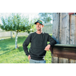 Un homme portant un sweat-shirt gris foncé John Deere avec un logo en relief et une casquette John Deere sourit en s’appuyant contre un mur en bois.