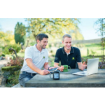 Two men wearing white and black John Deere polo shirts, one holding a green John Deere mug, at an outdoor table.