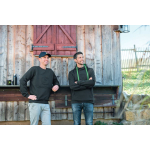 Deux hommes portent des vêtements John Deere : l’un avec un sweat-shirt noir à col rond et une casquette, l’autre avec un sweat à capuche noir et des cordons verts.