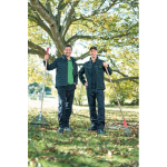 Dos hombres con ropa de trabajo oscura de John Deere, uno de ellos con una gorra, sonríen con rastrillos sobre un césped con hojas otoñales.