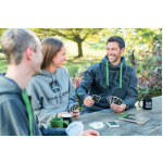 Une femme portant un sweat à capuche gris John Deere et deux hommes portant des sweats à capuche gris foncé John Deere avec des cordons verts jouent aux cartes en plein air.