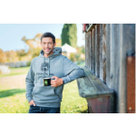 A smiling man in a grey John Deere hoodie holds a black John Deere mug while leaning against a wooden wall.