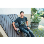A man wearing a dark t-shirt with a John Deere machinery graphic and dark jeans sits on a bench, holding a grey and black John Deere cap.