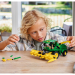 A young boy builds a green and yellow LEGO Technic John Deere combine harvester on a wooden table.